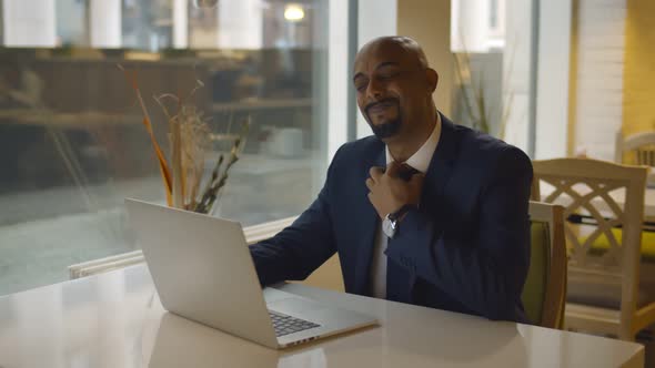 African Mature Businessman Loosening Tie Sitting at Table in Cafe and Working on Laptop alt