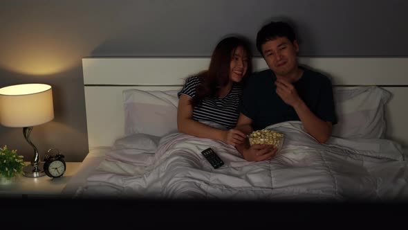 young couple watching TV and eating popcorn on a bed at night