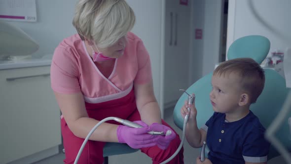 Dentist in Medical Mask and Gloves Ready To Checking Tooths of Little Carefree Boy Sitting alt
