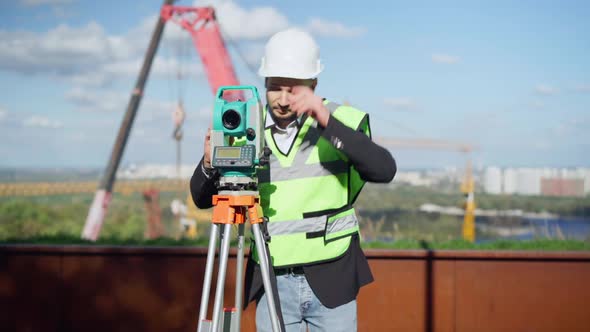Portrait of Positive Professional Male Engineer Using Theodolite Outdoors Taking Off Hard Hat alt