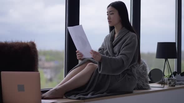 Side View Wide Shot of Concentrated Asian Slim Young Beautiful Woman in Bathrobe Sitting on alt