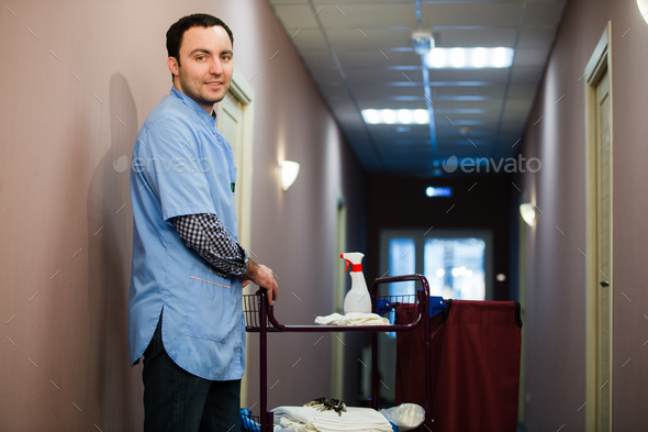 A man who is on the hotel cleaning crew staff is smiling with a towel vacuum in the process of