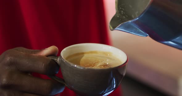 Midsection of african american barista pouring milk from jar into coffee cup in cafe alt