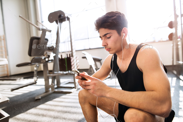 Hispanic man in gym resting, holding smart phone, listening music