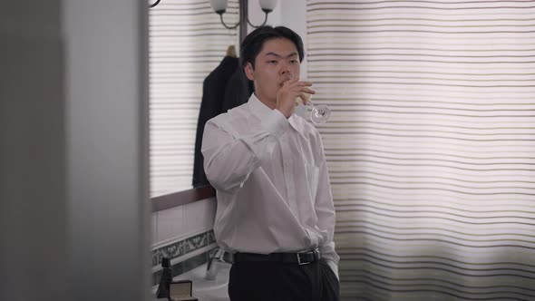 Portrait of Confident Asian Groom Drinking Champagne Standing in Bathroom in the Morning alt