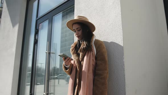 The Girl Stands Near a White Wall and Uses a Mobile Smartphone alt