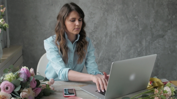 Female Florist Talking on Mobile Phone While Using Laptop in Flower Shop, Check Order Online alt