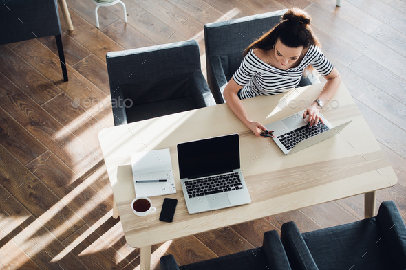 Multitasking woman in a cafe, working on both phone and laptop, while her coworker is resting. Stock Photo by romankosolapov