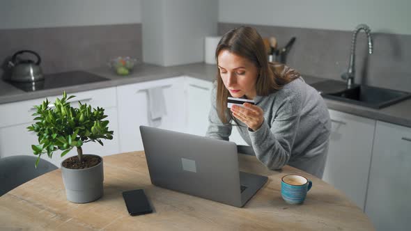 Woman Standing in Home Cozy Kitchen Leaning Over the Table and Makes an Online Shopping Using a alt