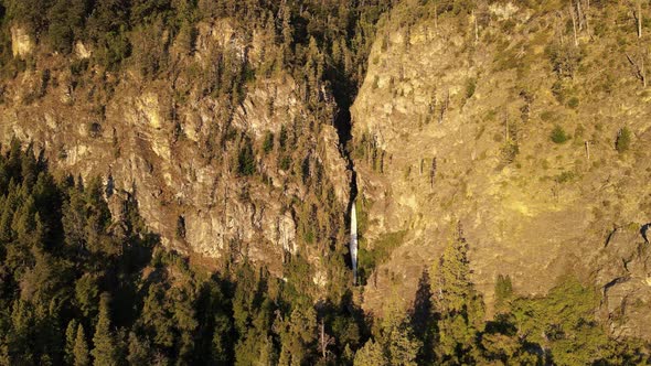 Aerial lowering over Corbata Blanca waterfall hiding between steep mountains with pine trees at suns alt