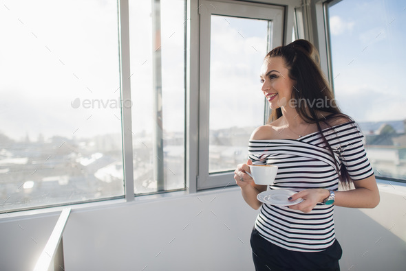 Happy woman in striped blouse smiling and standing indoors with a cup and looking away through the