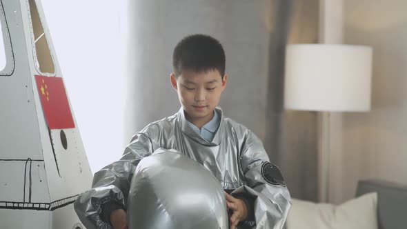 An Asian Boy in an Astronaut Costume Stands Near a Cardboard Model of a Spaceship and Puts on Helmet alt
