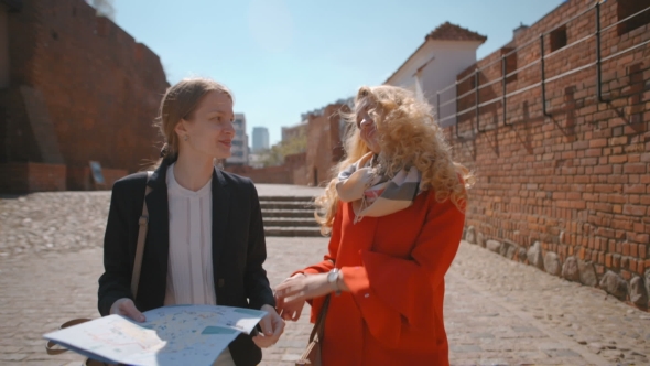 Two Women Tourists Are Smiling and Talking While Walking in Street with Map alt