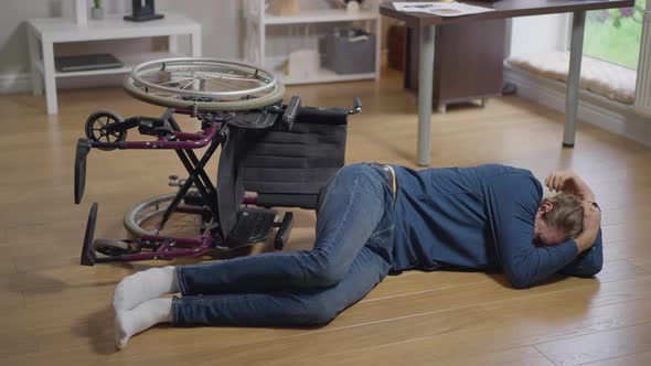 Wide Shot Crying Disabled Man Lying on Floor with Turned Over Wheelchair alt