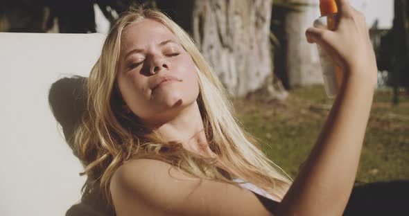 Woman Closing Her Eyes To Spray Sunscreen On Face alt