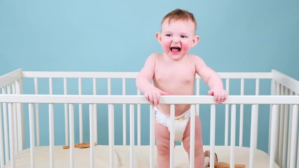 Naughty infant baby boy stands in the crib, studio blue background. Crybaby child in white diaper alt