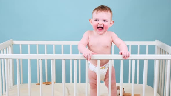 Naughty infant baby boy stands in the crib, studio blue background. Crybaby child in white diaper