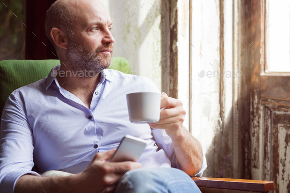 A man drinks coffee and checks the news in the phone, sitting in a chair by the window Stock Photo by koldunov