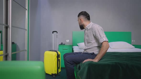 Side View of Happy Bearded Male Tourist Sitting on Bed in Green Hotel Room Smiling alt