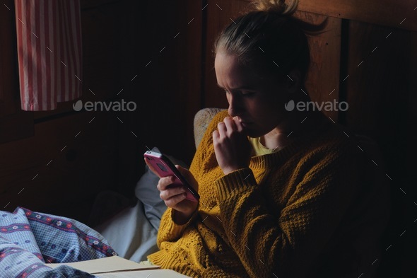 woman using laptop/smartphone in bed by window, rustic wooden cottage interior