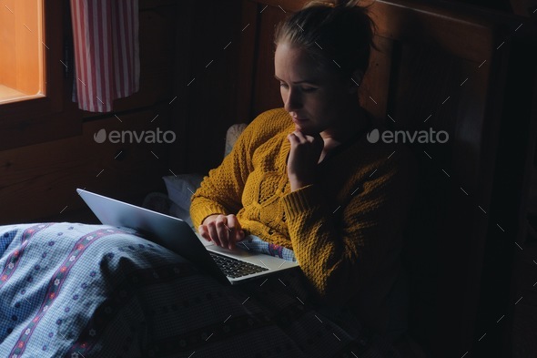 woman using laptop/smartphone in bed by window, rustic wooden cottage interior