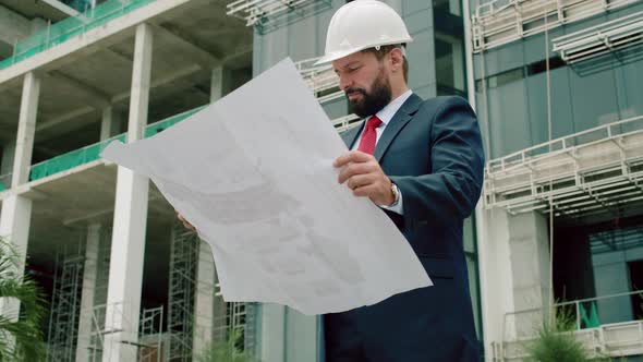 Engineer Male in a White Safety Helmet Developer at a Construction Site Checks on the Drawings at alt
