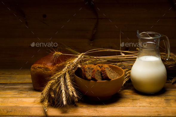 Glass jug with milk, mug with milk, a loaf of rye bread, ears on the background of wooden boards