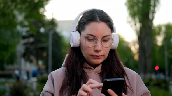 Beautiful Girl with Glasses and White Headphones Uses a Smartphone on the Street alt