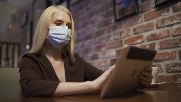 Attractive Young Woman in a Medical Mask Scrolls the Tablet in a Cafe alt