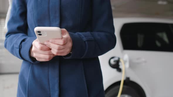 Woman Using Her Mobile Phone on the Background of the Charging Electric Car at the Parking alt