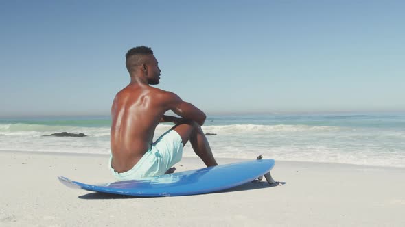 African American man sitting at beach with surfboard alt