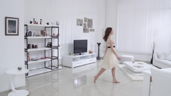Beautiful Young Woman with Bare Feet and a Beige Dress Moves in the Interior in the Living Room  alt