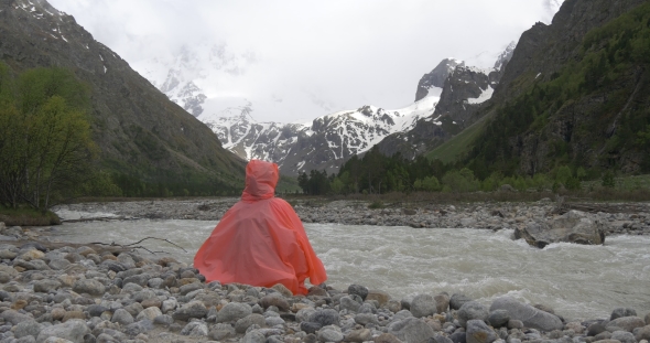 Person in Raincoat Sitting Near Mountain River
