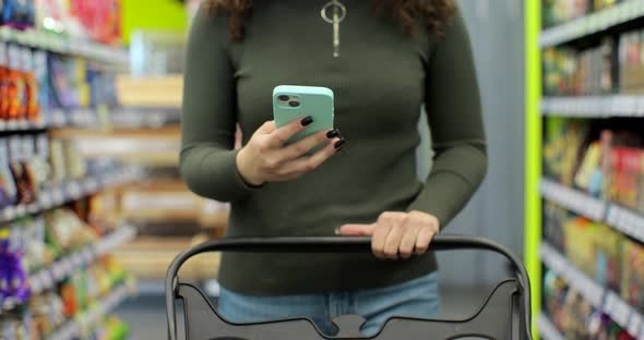 Close Up of Unrecognizable Woman Using Smartphone While Carrying Shopping Cart Walking Through alt