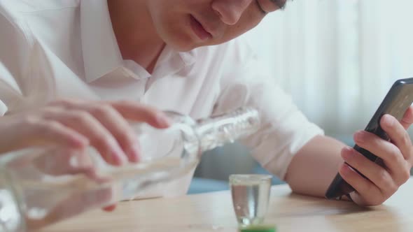 Close Up Of Drunk, Depressed Asian Man Pouring Vodka Before Drinking While Using Smartphone alt