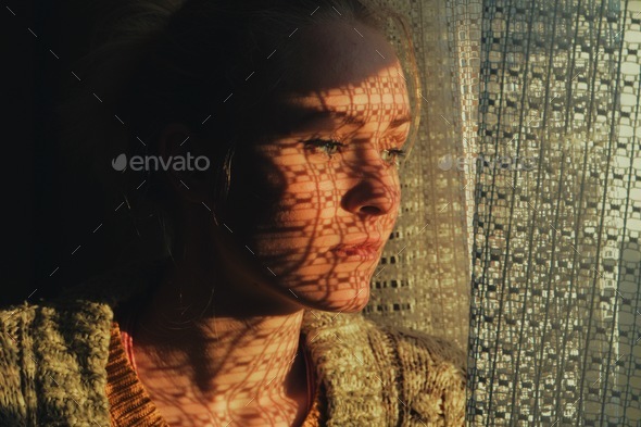portrait of a woman standing next to a window, sunset through curtain cast a shadow on her face