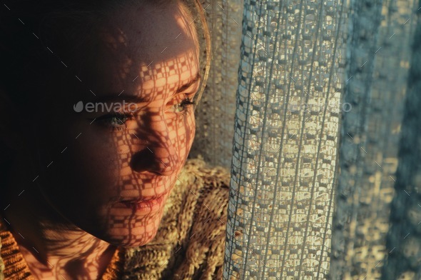 portrait of a woman standing next to a window, sunset through curtain cast a shadow on her face