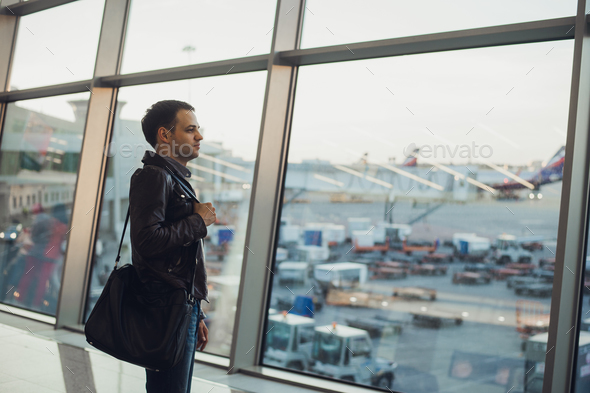 Travel concept with young man in airport interior with city view and a plane flying by. Stock Photo by romankosolapov