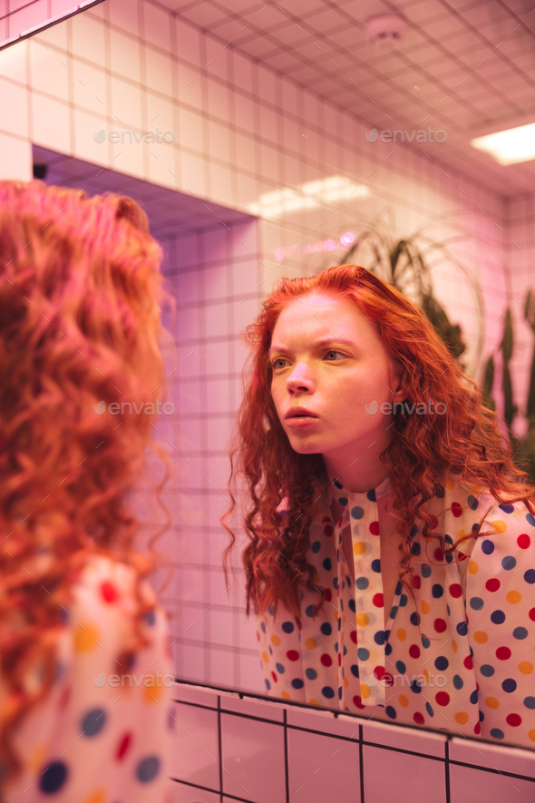 Concentrated young redhead curly lady looking at mirror.