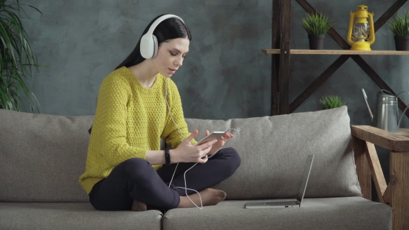 Young Woman Student in Headphones Using Smartphone on Sofa at Home