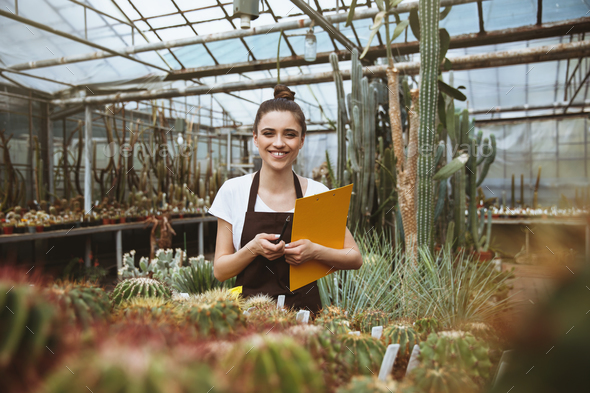 Happy young woman standing in greenhouse holding clipboard.
