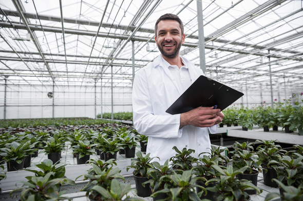 Happy man standing in greenhouse near plants holding clipboard