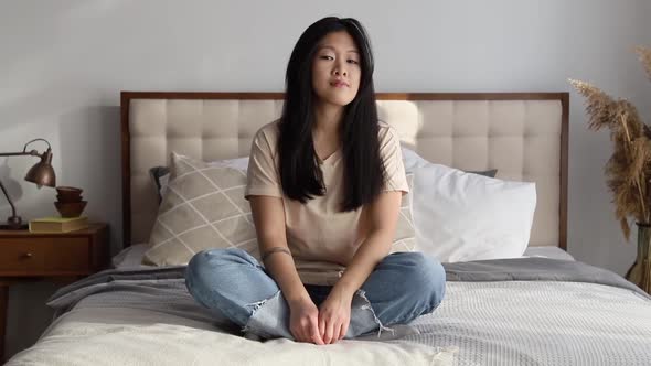 Attractive Smiling Asian Woman in Casual Clothes Sitting on the Bed at Home alt