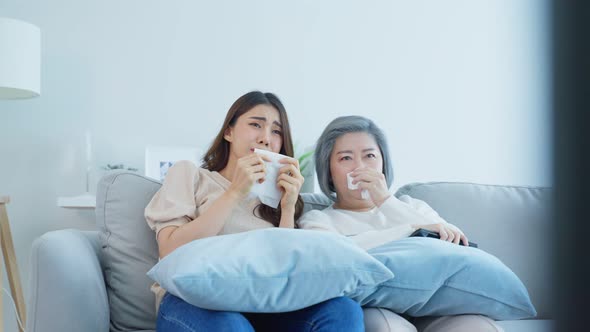 Asian senior mature woman and daughter sitting on sofa watch sad movie on TV and cry together. alt