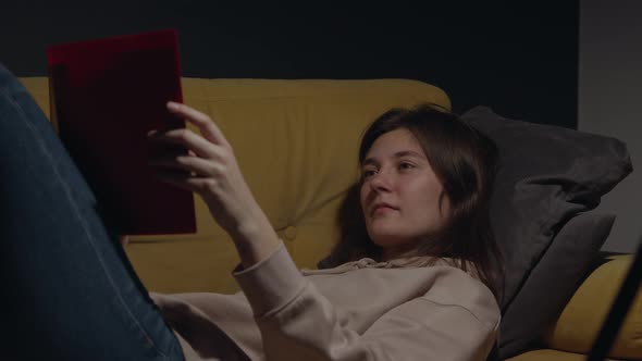 Attractive Girl Lies at Home on Sofa Reading Book Yawning Going to Sleep