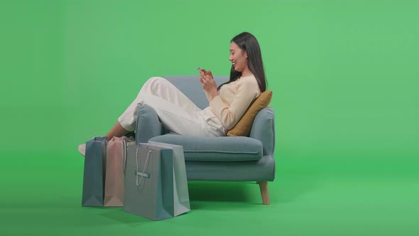 Woman With Shopping Bags Using Mobile Phone Shopping Online While Sitting On Sofa At Green Screen alt