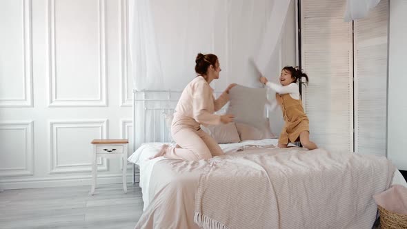 Happy Family Young Mom Baby Sitter and Little Kid Daughter Enjoy Funny Pillow Fight on Bed Carefree