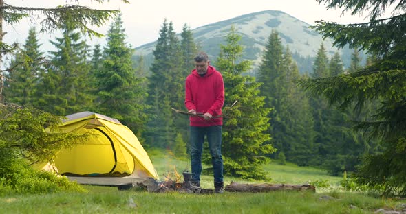 A Bearded Man Kindles a Fire on a Beautiful Forest Lawn in the Mountains alt