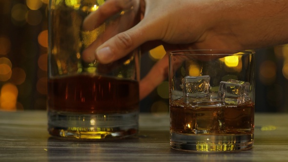 Pouring of Golden Whiskey, Cognac or Brandy From Bottle Into Glass with Ice Cubes. Shiny Background alt