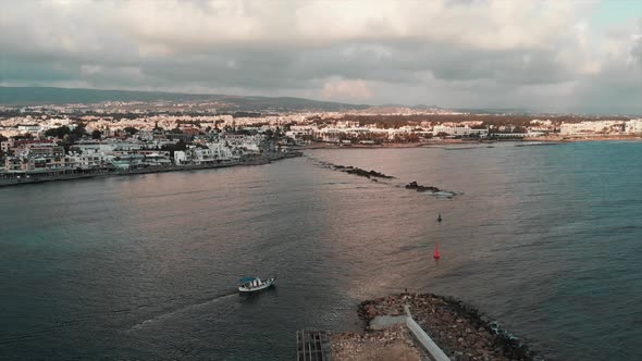 Small white fishing boat with blue roof sails into open sea at sunset in industrial marine city   alt